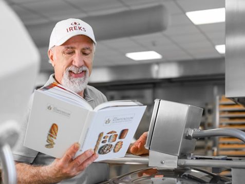 Ein Bäcker steht in der Bäckerei und liest im IREKS-Kompendium der Bäckereitechnologie